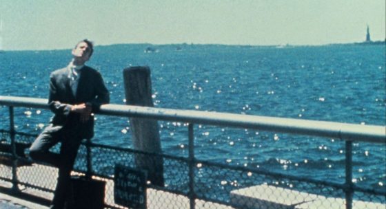 Mann in dunklem Anzug lehnt an einem Geländer mit Blick auf das glitzernde Meer, im Hintergrund die Freiheitsstatue.