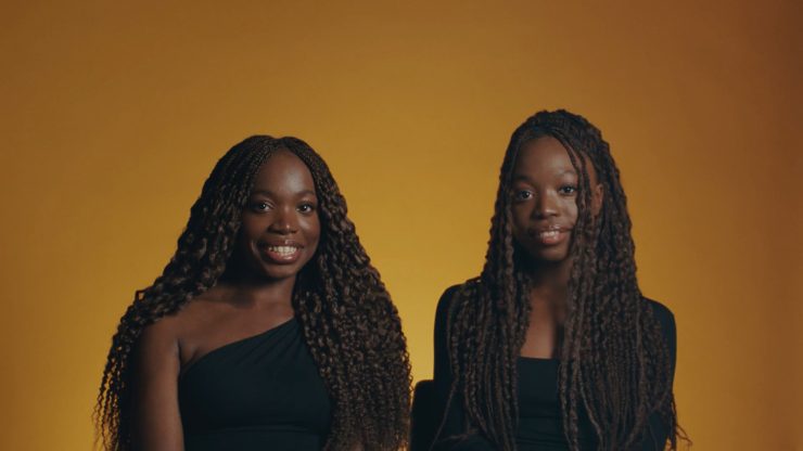 Zwei junge Frauen sitzen nebeneinander und blicken in die Kamera. Hinter ihnen ein oranger Hintergund.