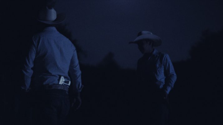 Zwei Männer in Cowboyhüten stehen nachts im Freien. Die Szene ist dunkel und in blauen Farbtönen gehalten, nur schwach vom Mondlicht beleuchtet. Der Mann links trägt ein weißes Holster am Gürtel, beide tragen Hemden und Jeans. Im Hintergrund sind schemenhaft Bäume zu erkennen.