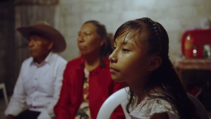 Ein junges Mädchen sitzt im Vordergrund, zwei ältere Erwachsene sitzen hinter ihr in einem schwach beleuchteten Raum mit kahlen Wänden.
