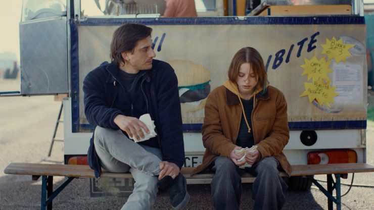 Eine weibliche junge Figur und eine männliche ältere sitzen vor einem Imbissstand und unterhalten sich.