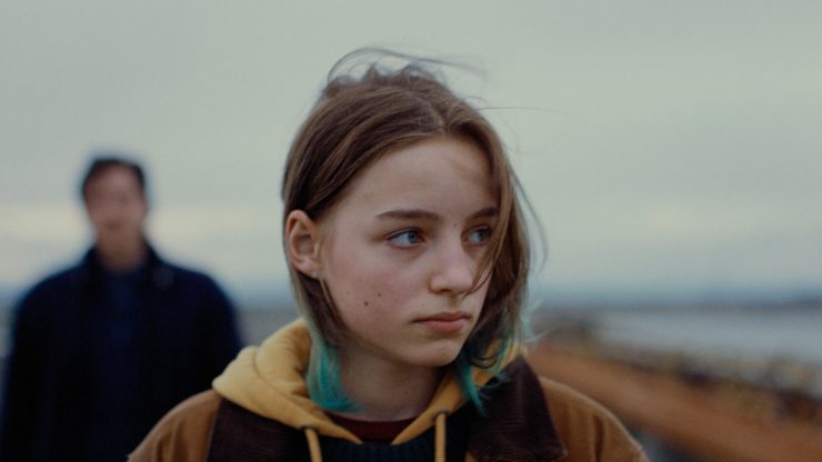 Filmstill: Weibliche Figur blickt zur Seite, im Hintergrund eine verschwommene Gestalt - herbstliche Umgebung am Meer.