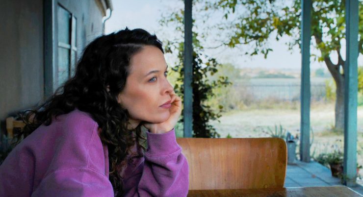 Filmstill aus 'MiroirsNo3': Eine Frau starrt nachdenklich in die Ferne - ländliche Ungembung mit einem See und Haus