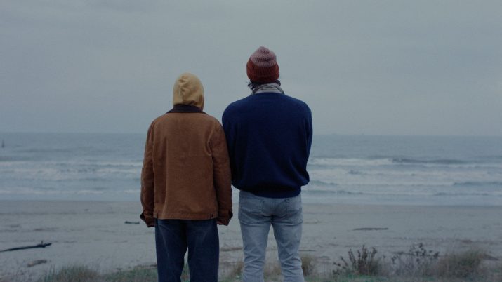 Zwei Personen von hinten, eine trägt eine braune Jacke und eine beige Kapuze, die andere eine blaue Jacke und eine rote Mütze, sie blicken auf das Meer bei bewölktem Himmel.