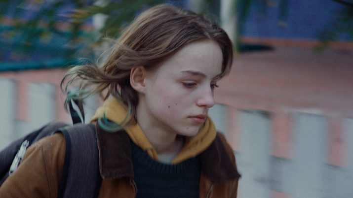 Ein junges Mädchen mit braunen Haaren trägt gelbe Kapuzenjacke unter braunem Mantel und schwarzen Rucksack und geht schnell, verschwommener Hintergrund.