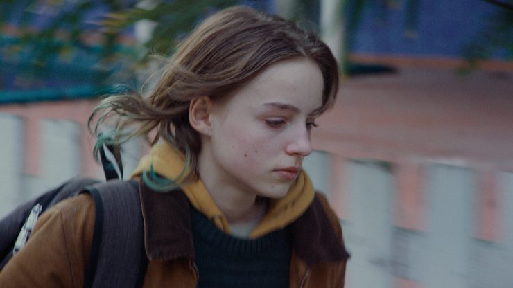 Ein junges Mädchen mit braunen Haaren trägt gelbe Kapuzenjacke unter braunem Mantel und schwarzen Rucksack und geht schnell, verschwommener Hintergrund.