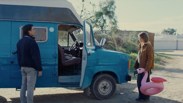Blauer Van mit geöffneter Fahrertür, zwei Personen stehen davor, eine trägt eine pinke Flamingo-Schwimmhilfe, draußen an einem sonnigen Tag.