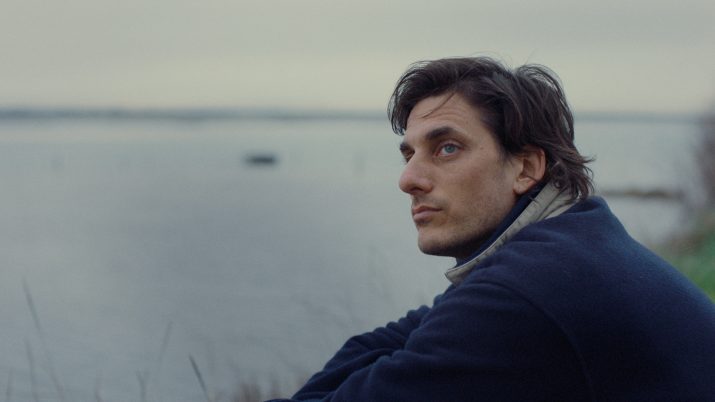 Person mit dunklem, gewelltem Haar trägt einen dunkelblauen Mantel und einen hellen Schal, sitzt seitlich am Ufer eines Gewässers.