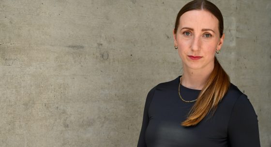 Frau mit langem braunem Pferdeschwanz, trägt schwarzes langärmeliges Oberteil und goldene Kette, steht vor Betonwand