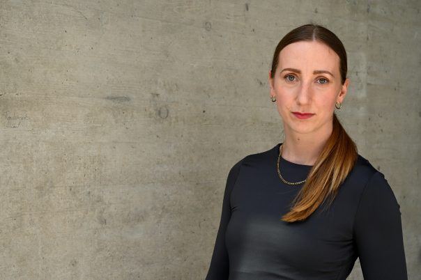 Frau mit langem braunem Pferdeschwanz, trägt schwarzes langärmeliges Oberteil und goldene Kette, steht vor Betonwand