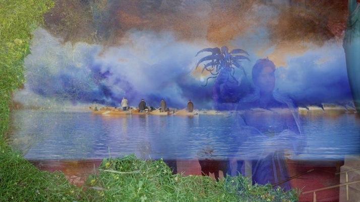 Sechs Personen sitzen am Ufer eines Gewässers, im Hintergrund eine große, mehrarmige Skulptur und neblige, farbige Wolken