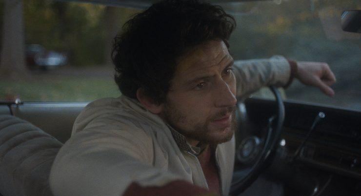 Filmstill: Ein Mann sitzt am Lenkrad im Auto, die Kamera blickt auf ihn vom Rücksitz aus. Er stützt einen Arm auf das Lenkrad, den anderen Arm auf die Rücklehne.