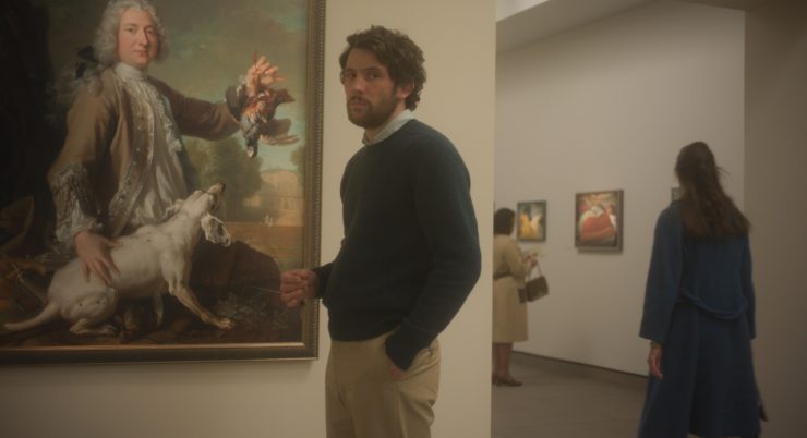 Filmstill: Ein Mann steht vor einem Gemälde in einer Ausstellung. Eine Hand in der Hosentasche, in der anderen hält er einen Schlüssel.