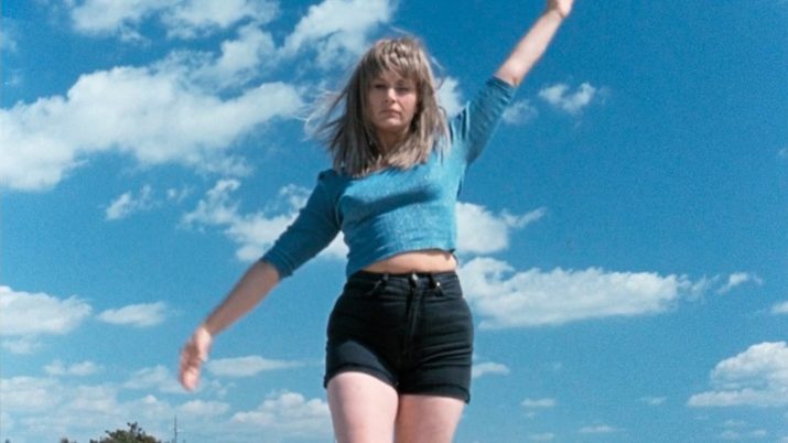 Frau mit blauem, langärmligem Oberteil und schwarzen Shorts springt mit erhobenen Armen vor blauem Himmel mit Wolken