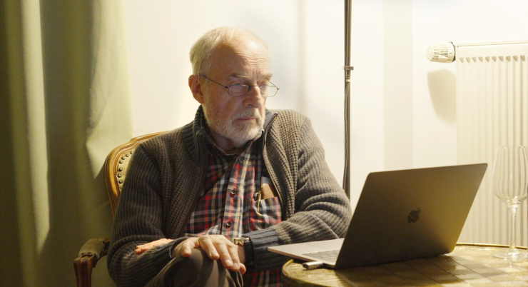 Älterer Mann mit grauem Strickpullover und kariertem Hemd sitzt auf einem Stuhl neben einem runden Holztisch mit einem geöffneten MacBook und einem leeren Weinglas.