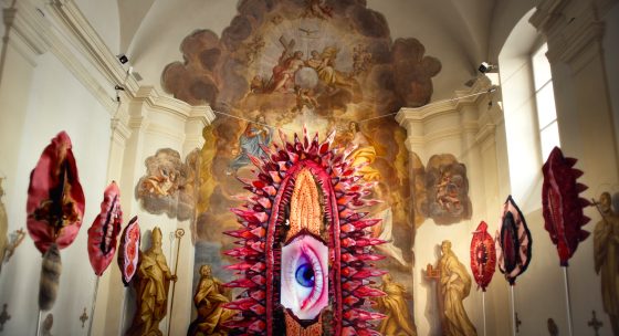 Der Innenraum einer Kirche. An den Wänden sind künstlerisch gestaltete Vulven aufgehängt. In der Mitte ein großes Auge.
