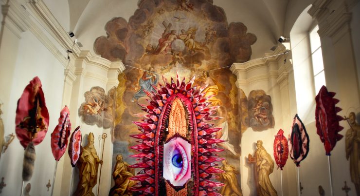 Der Innenraum einer Kirche. An den Wänden sind künstlerisch gestaltete Vulven aufgehängt. In der Mitte ein großes Auge.