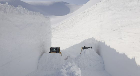 Zwei Schneefräsen räumen einen schmalen Weg zwischen hohen Schneewänden in einer verschneiten Berglandschaft.
