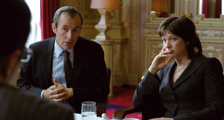 Zwei Personen in formeller Kleidung sitzen an einem Tisch in einem elegant eingerichteten Raum mit rotem Teppich und goldfarbenen Wänden.