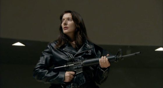 Person mit langen dunklen Haaren trägt schwarze Lederjacke und hält ein Gewehr in beiden Händen.