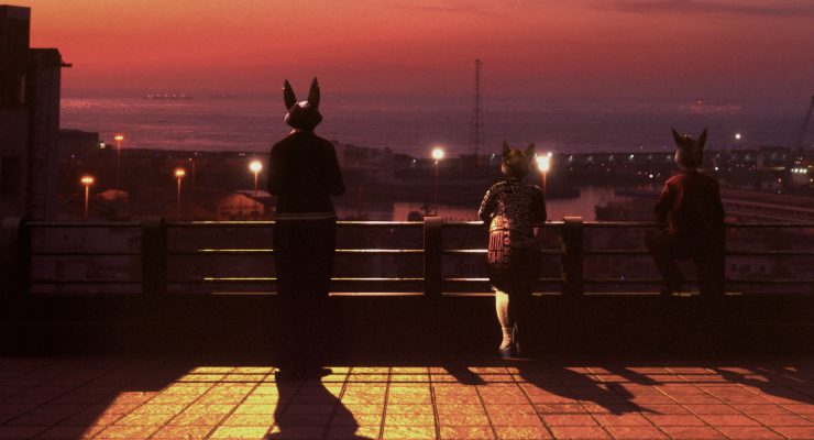 Drei Personen mit Tiermasken stehen auf einer Terrasse und blicken auf eine beleuchtete Stadt bei Sonnenuntergang.