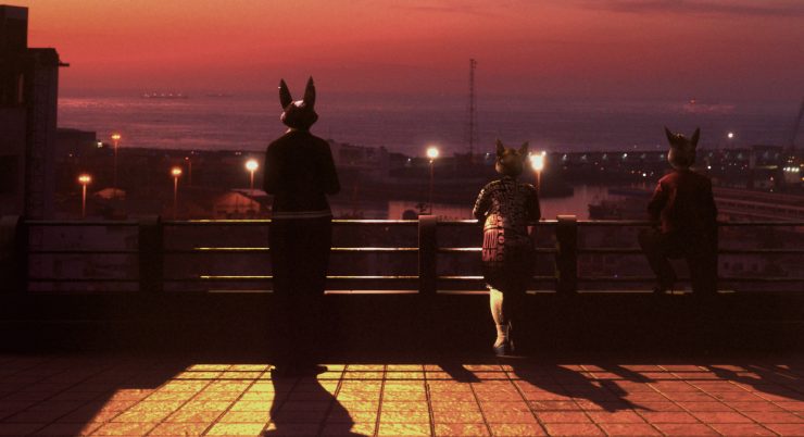 Drei Personen mit Tiermasken stehen auf einer Terrasse und blicken auf eine beleuchtete Stadt bei Sonnenuntergang.