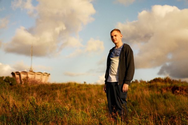Person steht auf einer Wiese mit langem Gras, trägt schwarze Jacke, weißes T-Shirt und dunkle Hose, im Hintergrund Wolken und ein Bauwerk.