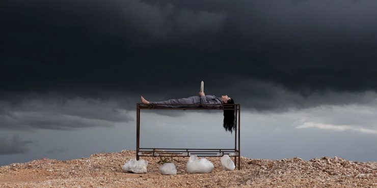 Person liegt auf dem Rücken auf einem Metallgestell, dunkle Wolken am Himmel, felsiger Boden mit weißen Steinen