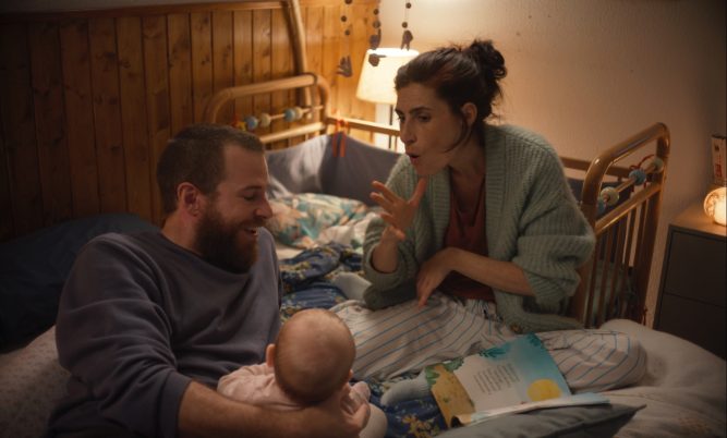 Zwei Erwachsene sitzen auf einem Bett, eine Person hält ein Baby, eine andere gestikuliert mit der Hand. Im Hintergrund Holzvertäfelung und eine Lampe.