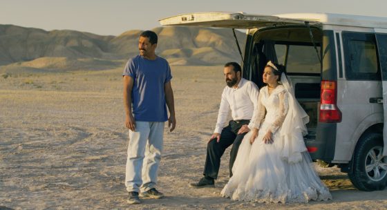 Brautpaar in weißem Hochzeitskleid und schwarzem Anzug sitzt am geöffneten Heck eines weißen Vans in einer kargen Wüstenlandschaft, daneben steht ein Mann in blauem T-Shirt und heller Hose.