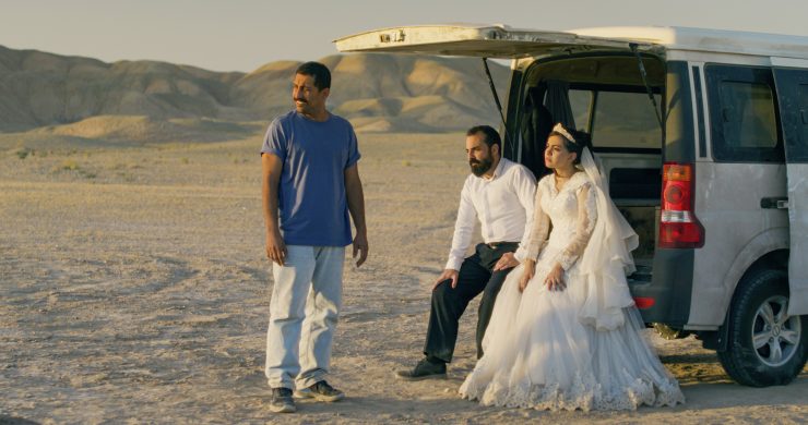 Brautpaar in weißem Hochzeitskleid und schwarzem Anzug sitzt am geöffneten Heck eines weißen Vans in einer kargen Wüstenlandschaft, daneben steht ein Mann in blauem T-Shirt und heller Hose.
