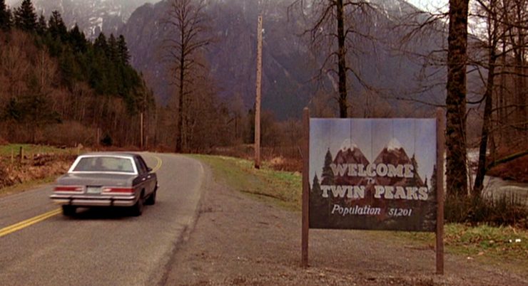 Ein Auto fährt auf einer kurvigen Landstraße, rechts steht ein Schild mit der Aufschrift 'WELCOME TWIN PEAKS Population 51,201'.