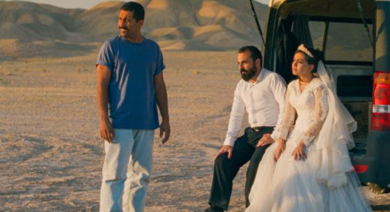 Drei Personen blicken nach links. Zwei von ihnen sitzen lehnen am Kofferraum eines Autos. Im Hintergrund Wüstenlandschaft.