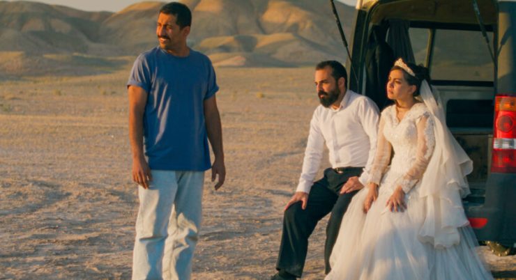 Drei Personen blicken nach links. Zwei von ihnen sitzen lehnen am Kofferraum eines Autos. Im Hintergrund Wüstenlandschaft.