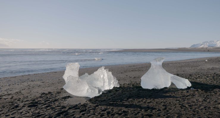 Zwei unregelmäßig geformte Eisstücke liegen auf einem dunklen Sandstrand, im Hintergrund das Meer und ein weiter Horizont.