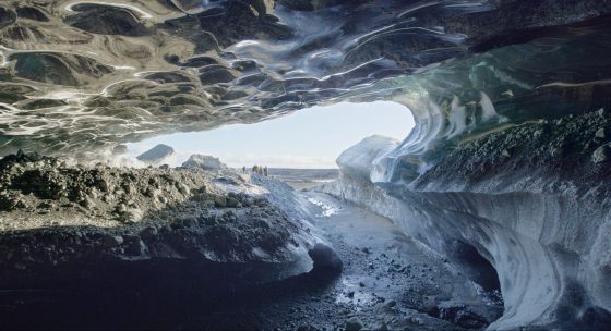 Blick aus einer Eishöhle mit glatten, gewölbten Eiswänden und mehreren Menschen am Höhleneingang im Hintergrund.