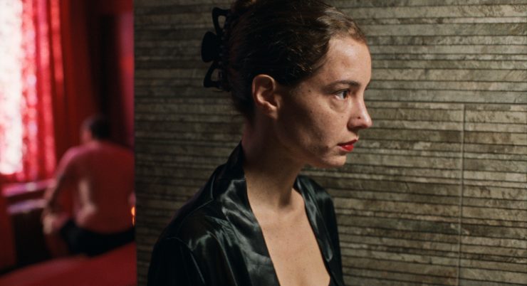 Eine junge Frau mit hochgesteckten Haaren blickt nach rechts. Hinter ihr sieht man einen Mann auf einer Bettkante sitzen.