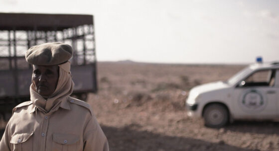 Filmstill: Eine Frau steht links im Bild, hinter ihr Wüste und ein weißes Auto