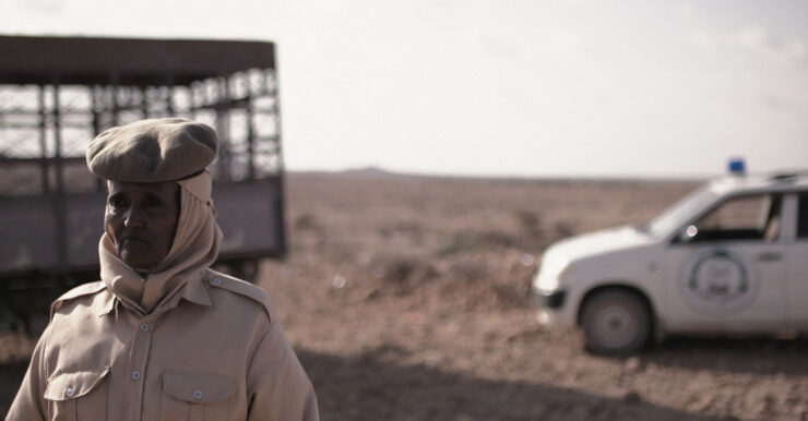 Filmstill: Eine Frau steht links im Bild, hinter ihr Wüste und ein weißes Auto