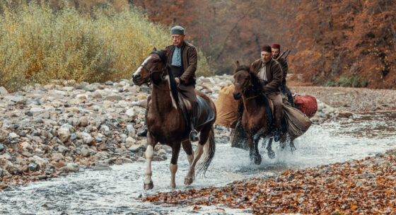 Drei Männer reiten auf Pferden durch einen flachen Fluss mit steinigem Ufer und herbstlich gefärbtem Laub im Hintergrund