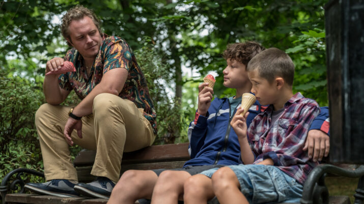 Drei Personen sitzen auf einer Parkbank, zwei Kinder essen Eis, ein Erwachsener sitzt mit angewinkeltem Arm daneben.