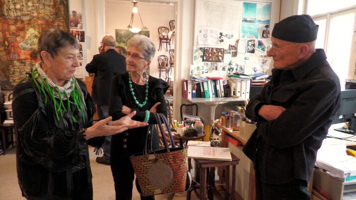 Drei Personen unterhalten sich in einem Raum mit Regalen, Tischen und zahlreichen Bildern an den Wänden