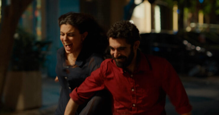 Zwei Personen rennen nebeneinander auf einer Straße in der Nacht, eine Person trägt ein rotes Hemd, die andere ein dunkles Oberteil