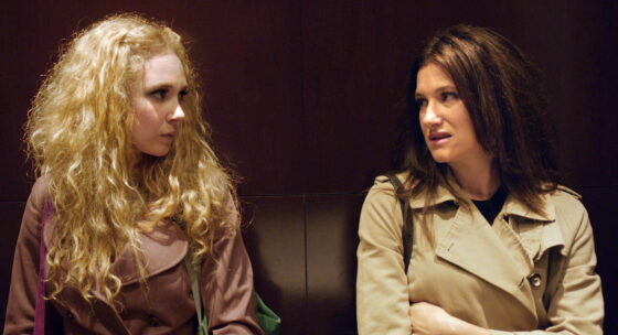 Zwei Frauen mit langen Haaren sitzen nebeneinander auf einer dunklen Ledercouch, eine trägt einen hellen Trenchcoat, die andere einen braunen Pullover.