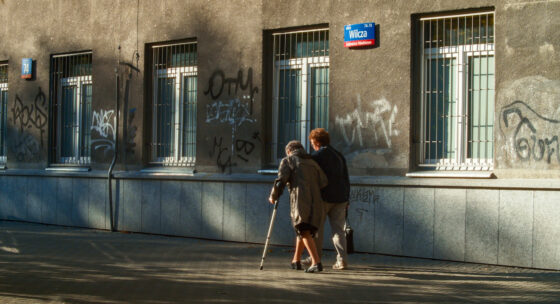 Zwei ältere Personen gehen an einer grauen Hauswand mit mehreren Fenstern und Graffiti entlang, eine Person stützt sich auf einen Gehstock
