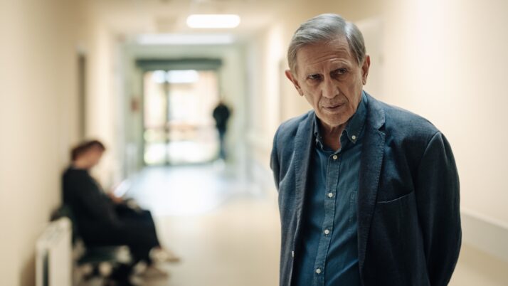 Älterer Mann mit grauen Haaren trägt dunkelblauen Blazer und hellblaues Hemd in einem hellen Flur mit Sitzbank und einer weiteren Person im Hintergrund.