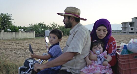 Mann mit Strohhut fährt Motorrad mit Kind auf dem Tank und Frau mit Baby auf dem Sozius vor offenem Feld und Gebäuden im Hintergrund