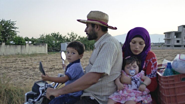 Mann mit Strohhut fährt Motorrad mit Kind auf dem Tank und Frau mit Baby auf dem Sozius vor offenem Feld und Gebäuden im Hintergrund