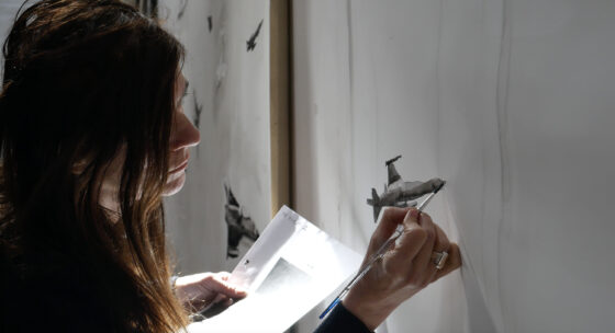 Frau mit dunklen Haaren hält Pinsel und Papier und malt an einer weißen Wand mit kleinen schwarzen Flugzeugzeichnungen