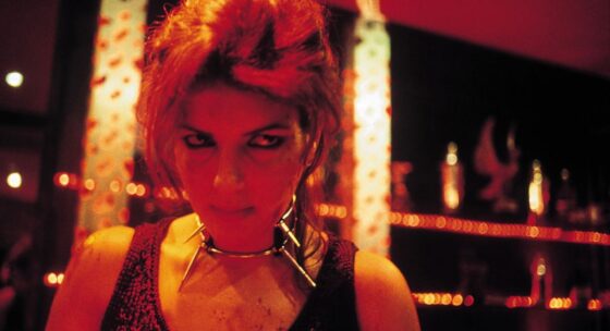 Frau mit hochgestecktem Haar und schwarzem ärmellosem Oberteil vor einer Wand mit Lichterketten und Dekorationen in warmem Licht